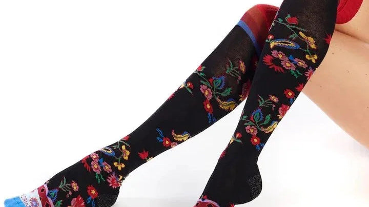 Black knee-high floral socks with colorful embroidery from Love Classic, worn by a seated model.