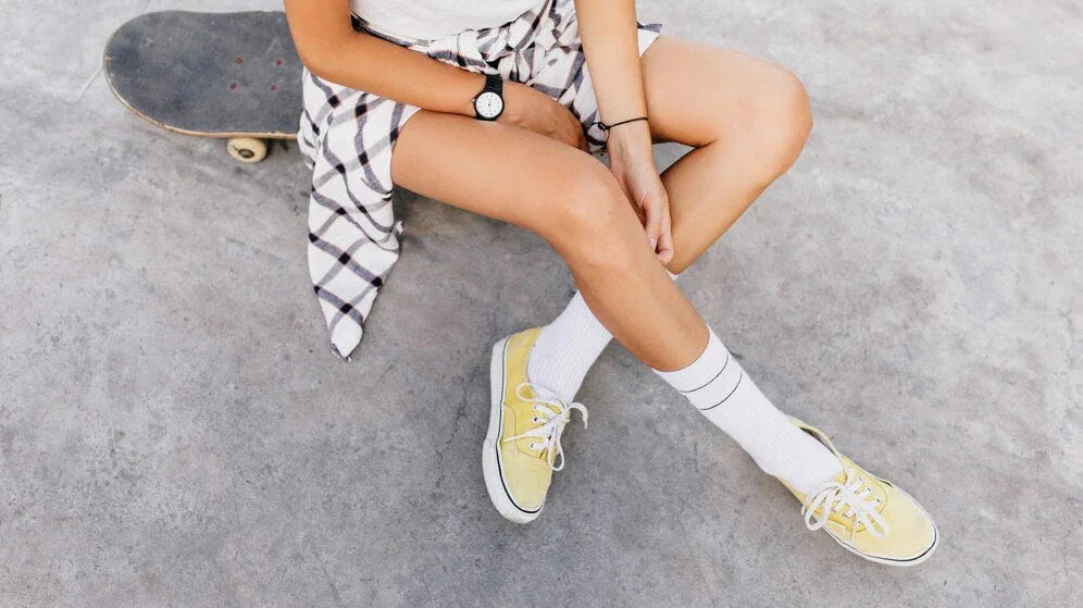 Woman in white socks and yellow sneakers sitting on skateboard at skate park, casual style