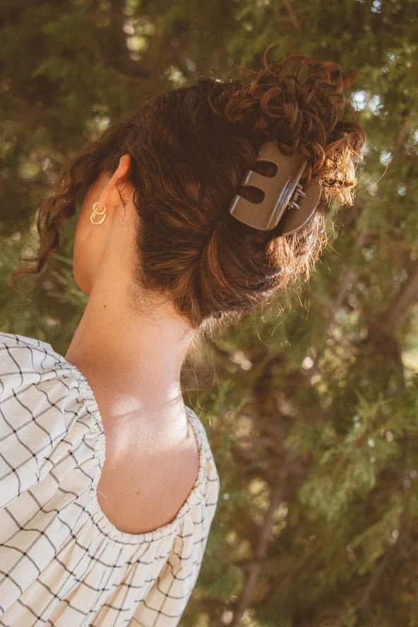Woman with curly hair in a large claw clip, wearing a checkered top, outdoors among trees