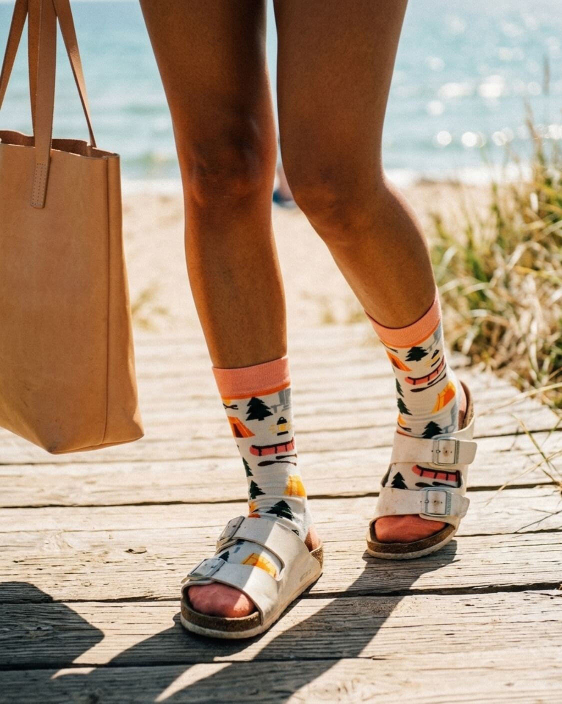 Crew Socks, Happy Camper featuring a colorful outdoor pattern with tents, trees, campfire graphics, and a soft ribbed cuff, worn with buckle sandals on a wooden beach walkway, highlighting playful casual styling for relaxed outdoor days.