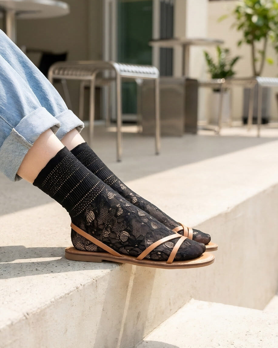 Sheer Floral Lace Crew Socks in black with delicate floral lace pattern, styled with rolled cuff jeans and strappy flat sandals while seated on outdoor concrete steps, highlighting the semi-transparent texture and breathable knit.