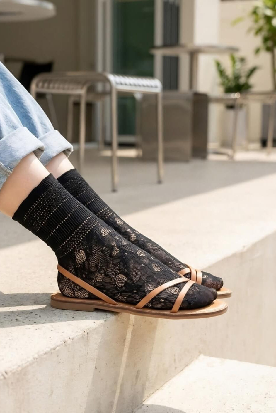 Sheer Floral Lace Crew Socks in black with delicate floral lace pattern, styled with rolled cuff jeans and strappy flat sandals while seated on outdoor concrete steps, highlighting the semi-transparent texture and breathable knit.