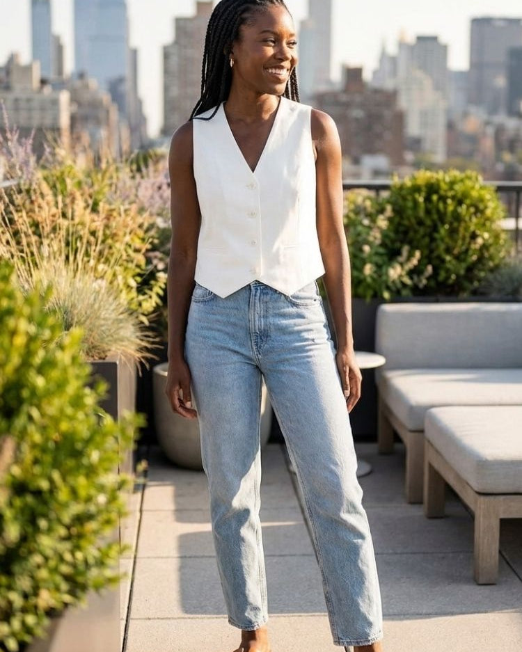 Naples Vest, sleeveless V-neck tailored vest in off-white with button front, styled with light-wash straight-leg jeans and tan sandals on a rooftop terrace, highlighting the clean structure and relaxed summer look.