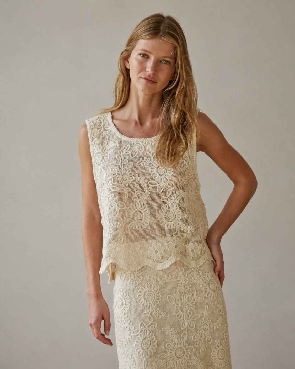 Sleeveless Embroidery Crop Top featuring intricate floral embroidery on a sheer mesh overlay in a soft ivory tone, styled with a scalloped hem and relaxed fit, worn by a model in a neutral studio setting, highlighting the delicate craftsmanship and lightweight elegance of the design.