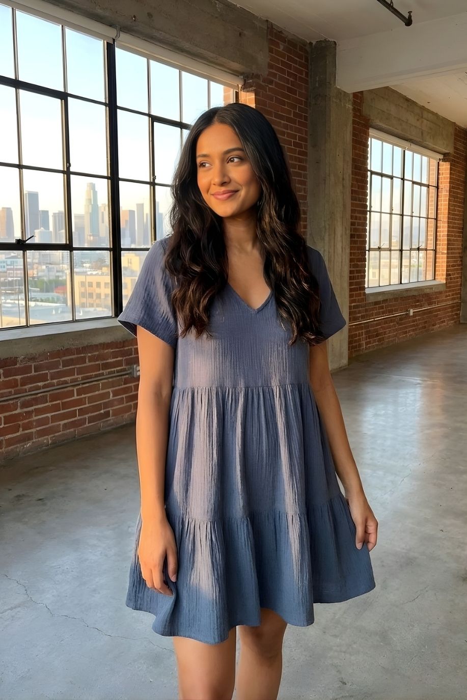 Cassady Dress featuring a relaxed, flowy silhouette with a V-neckline, short sleeves, and tiered skirt, styled indoors by large industrial windows with a city skyline view.