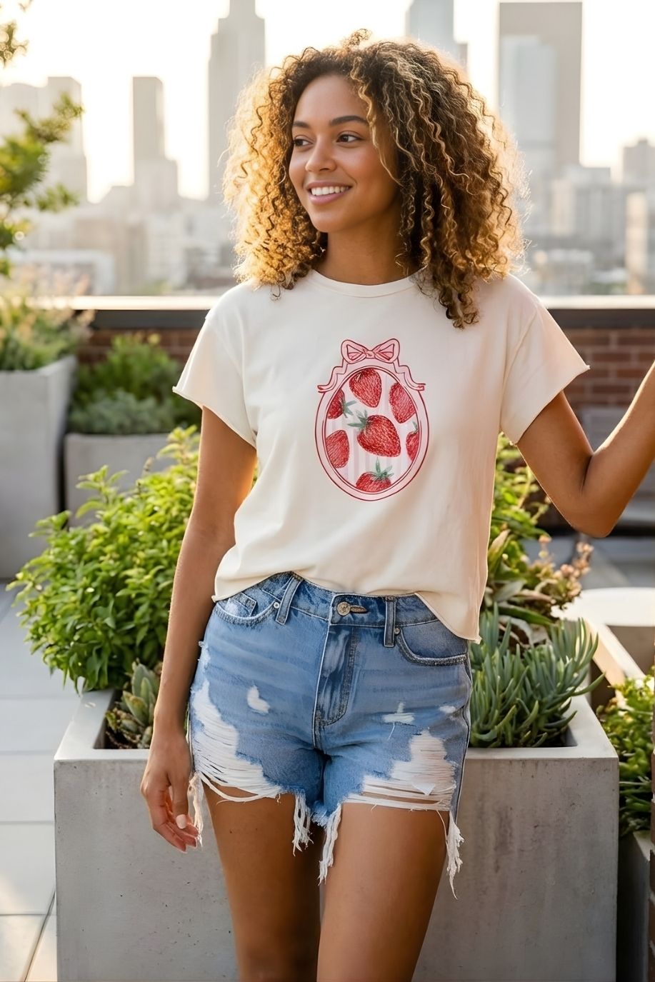 Strawberry Coquette American Vintage Graphic Crop Top featuring a cream short-sleeve design with red strawberry and bow artwork, styled with distressed denim shorts on a rooftop garden with city skyline in the background.