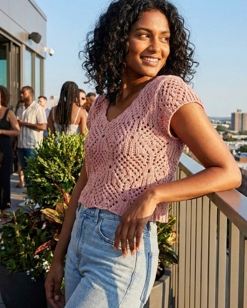 Crochet Knit Cap Sleeve Sweater Top, blush pink open-knit sweater with cap sleeves and scalloped V-neckline, styled with light-wash high-rise jeans, worn by a model leaning on a balcony railing at a rooftop gathering, highlighting the airy texture and relaxed fit in warm sunlight.
