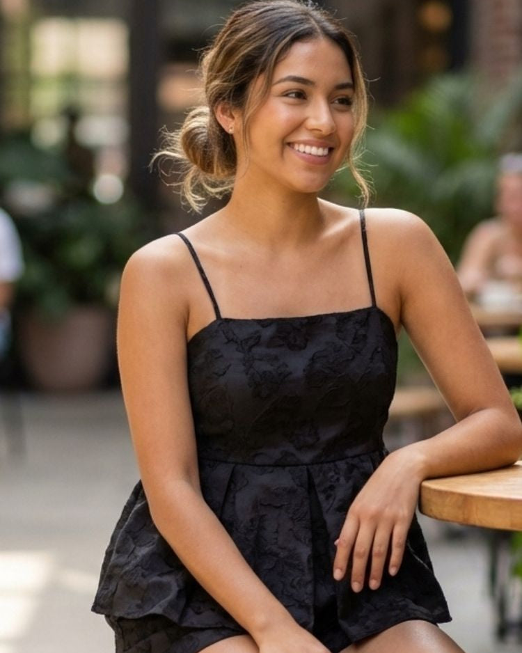 Sophie Top, black spaghetti strap peplum top with a tonal textured pattern, worn in a seated pose at a café table, emphasizing the structured bodice and soft volume at the waist.