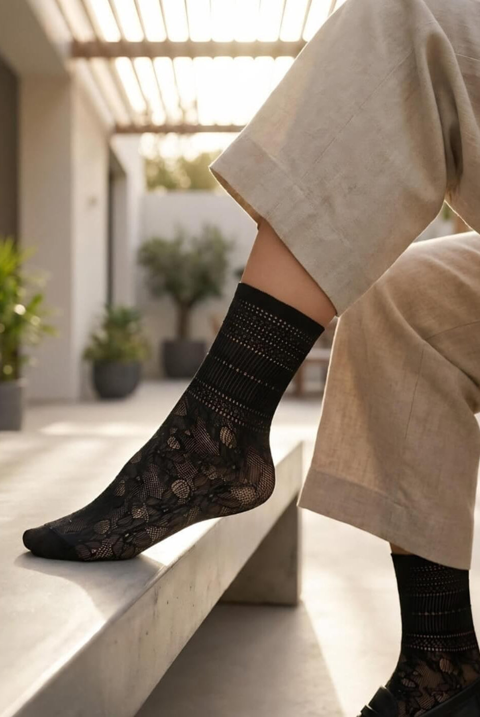 Sheer Floral Lace Crew Socks in black with intricate floral lace design, worn with tailored trousers and black loafers while sitting on a sunlit bench, showcasing the lightweight, sheer finish and elegant pattern.