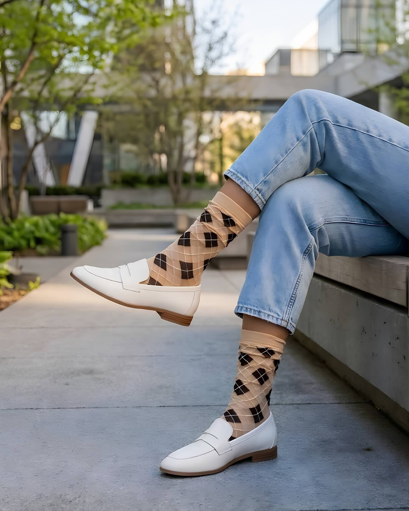 Sheer Argyle Crew Socks in beige and black featuring a soft diamond pattern, paired with white loafers and cuffed jeans while sitting on a concrete bench, showcasing the lightweight, sheer texture and casual styling.