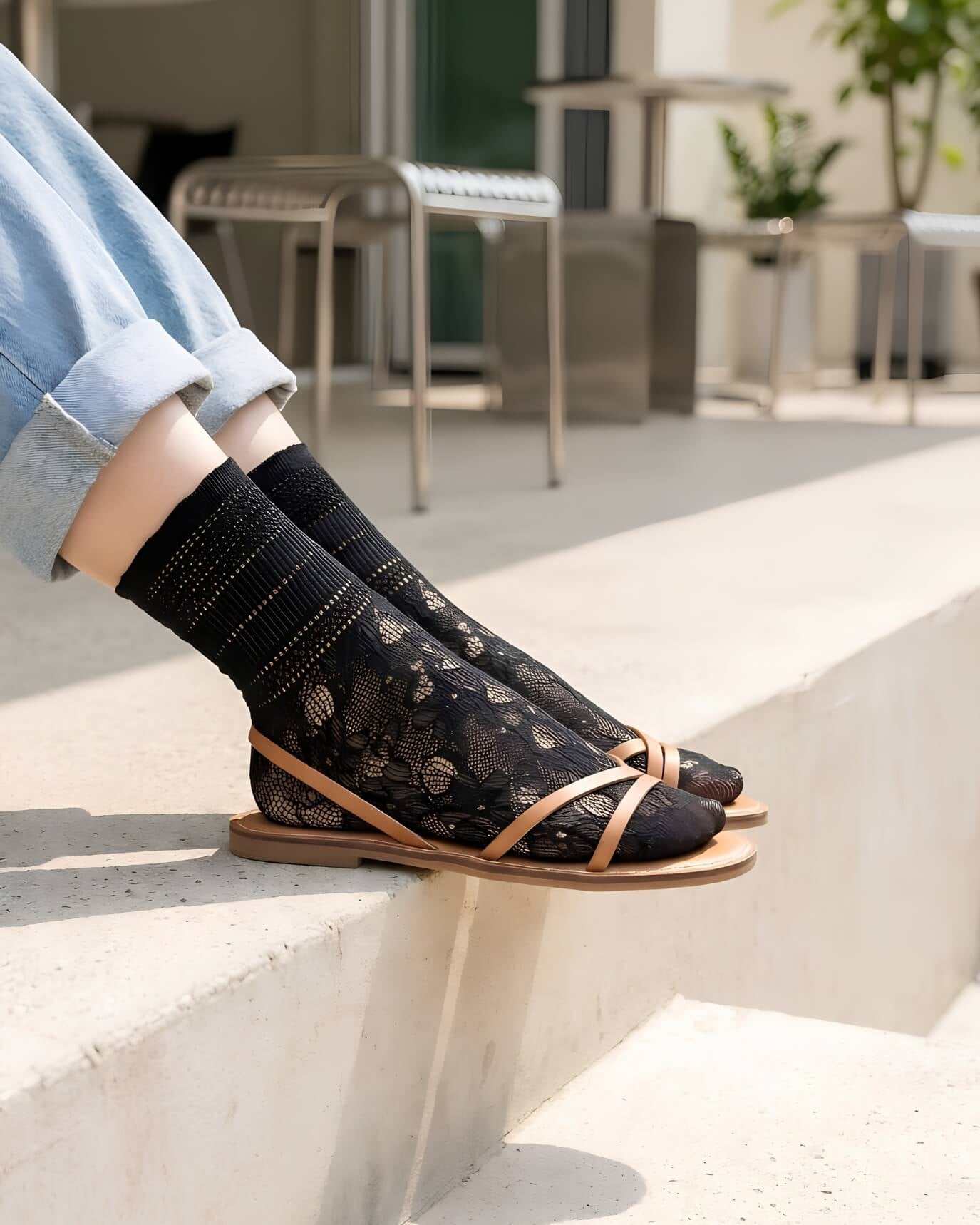 Sheer Floral Lace Crew Socks in black with delicate floral lace pattern, styled with rolled cuff jeans and strappy flat sandals while seated on outdoor concrete steps, highlighting the semi-transparent texture and breathable knit.