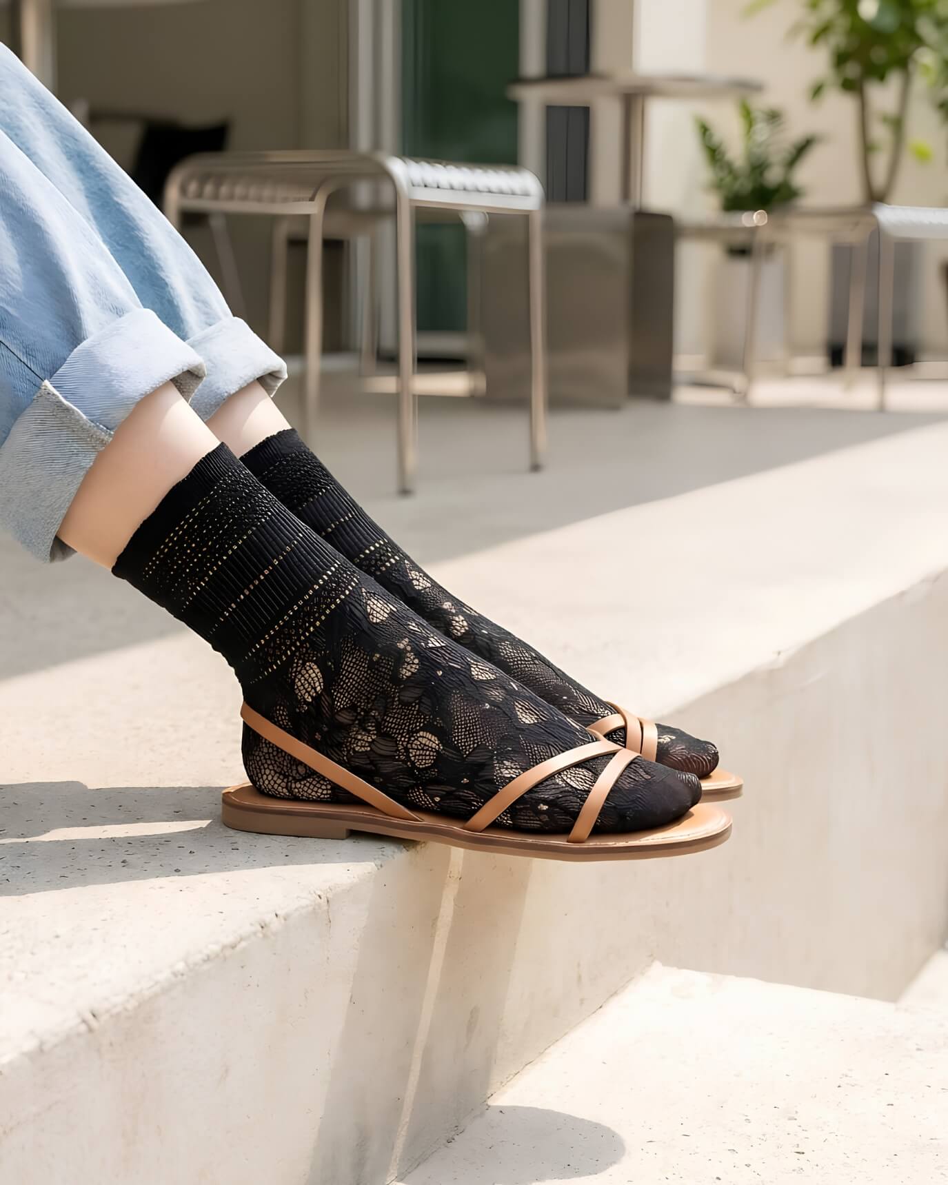 Sheer Floral Lace Crew Socks in black with delicate floral lace pattern, styled with rolled cuff jeans and strappy flat sandals while seated on outdoor concrete steps, highlighting the semi-transparent texture and breathable knit.