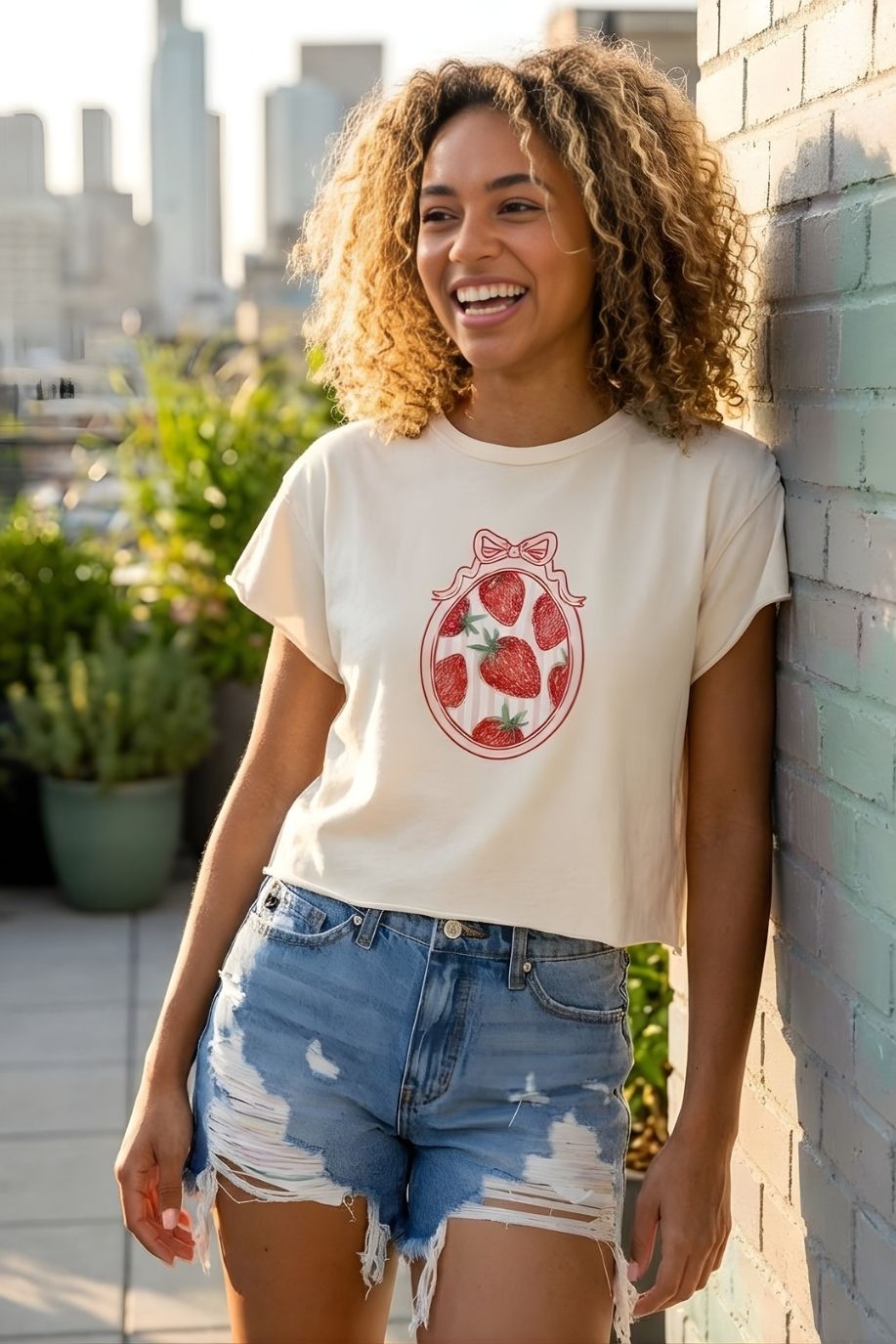 Strawberry Coquette American Vintage Graphic Crop Top featuring a relaxed cropped fit with vintage-inspired strawberry frame graphic, paired with ripped denim shorts and shown beside a sunlit brick wall.
