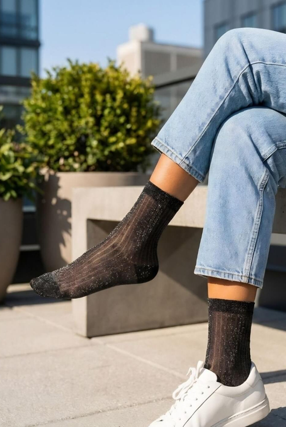 Sheer Metallic Ribbed Crew Socks in black with subtle shimmer, styled with light wash rolled cuff jeans and white sneakers while seated on a concrete bench, highlighting the semi-transparent ribbed texture and lightweight breathable fabric.
