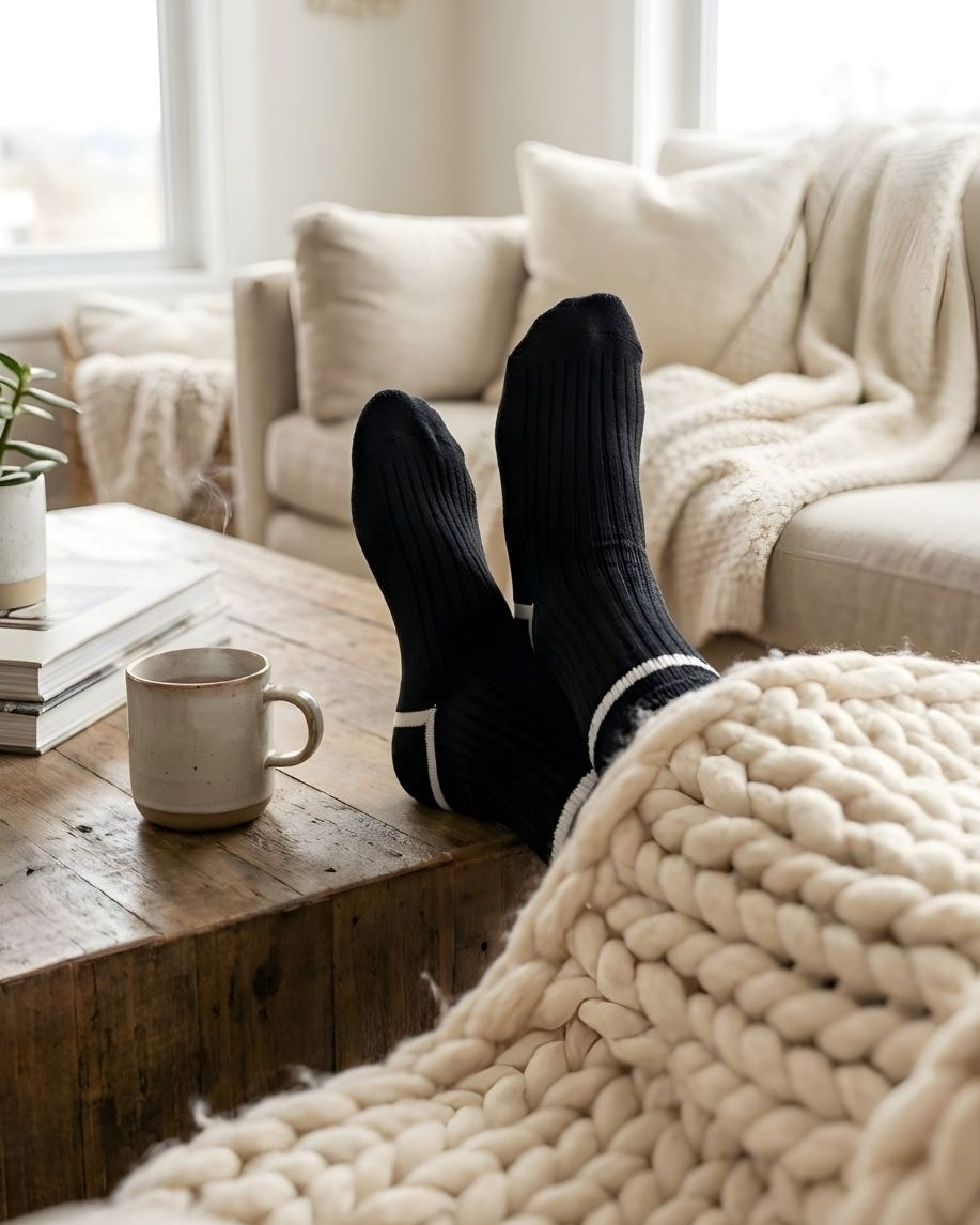 Classic Ribbed Crew Socks with Contrast Trim, black ribbed crew socks with white stripe accents worn while lounging on a sofa, styled with a cozy knit blanket and coffee table setting to evoke a relaxed, at-home feel.