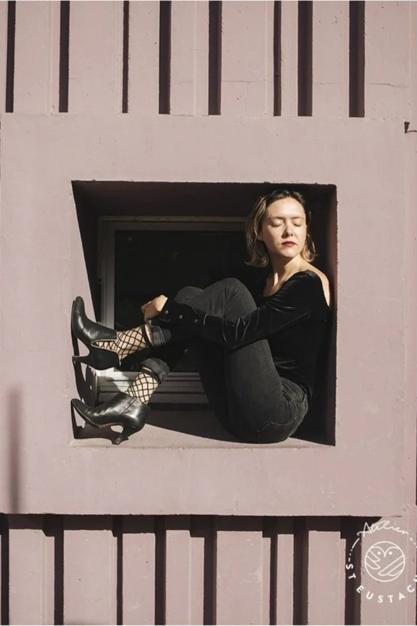 Woman in black velvet top, black pants, and heeled shoes sitting in a modern window, Love Classic style