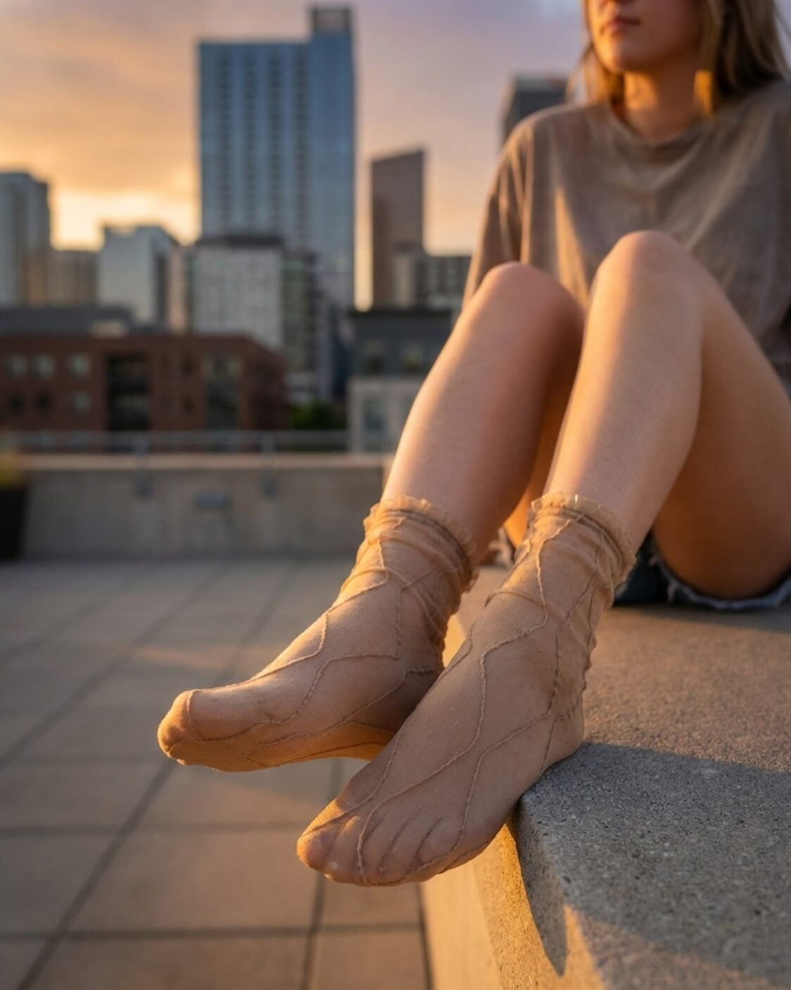 Bundled Packs Rosalie Lace Sheer Socks featuring nude sheer ankle socks with raised wavy lace texture and ruffled cuff trim, worn while seated on a rooftop ledge in warm sunset lighting, highlighting the lightweight translucent finish and delicate textured pattern.