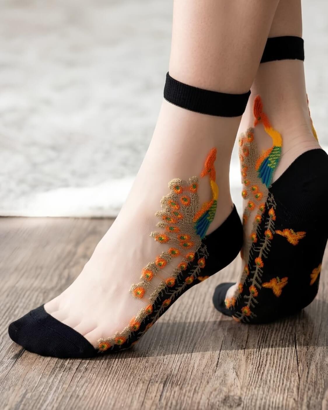 Peacock Sheer Mesh Socks featuring delicate nude-toned sheer mesh with colorful peacock embroidery and black reinforced toe, heel, and cuff trim, shown in a close-up standing pose on a wooden floor, emphasizing the lightweight texture, intricate stitching, and stylish feminine aesthetic.