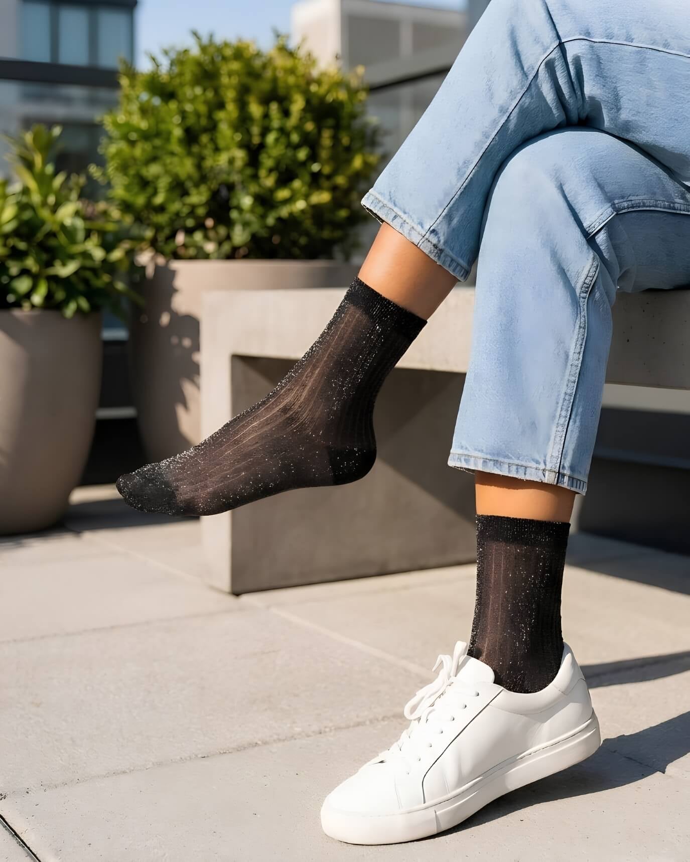 Sheer Metallic Ribbed Crew Socks in black with subtle shimmer, styled with light wash rolled cuff jeans and white sneakers while seated on a concrete bench, highlighting the semi-transparent ribbed texture and lightweight breathable fabric.