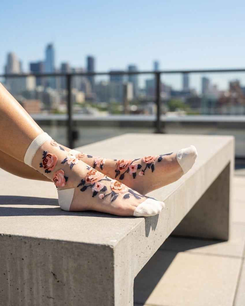 Roses Sheer Socks 2-Pack featuring sheer ankle socks with embroidered rose motifs and reinforced toe panels, worn while resting on a concrete bench in a bright city setting, highlighting the soft cuff fit and floral styling.