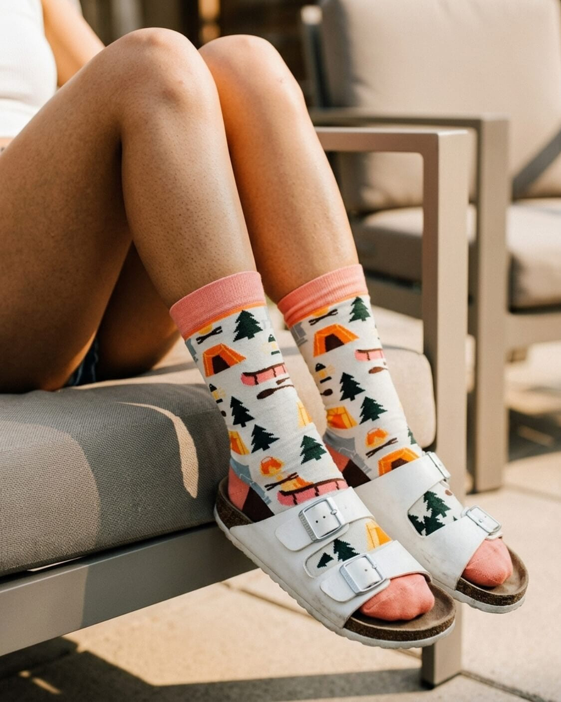 Crew Socks, Happy Camper featuring a full crew length with a pink cuff, camping graphics, and colorful outdoor motifs, worn with white buckle sandals while seated on patio furniture, highlighting the bold pattern and relaxed everyday wear.