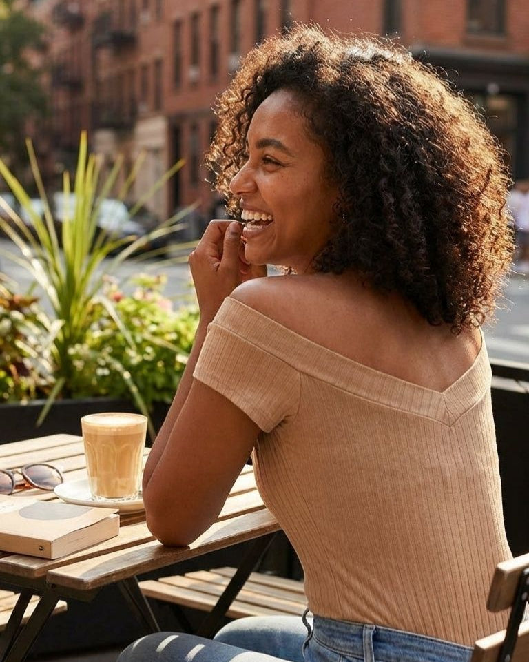 Larissa Top, back view showing the off shoulder neckline and fitted ribbed silhouette styled with denim while seated at an outdoor café table in warm natural light.