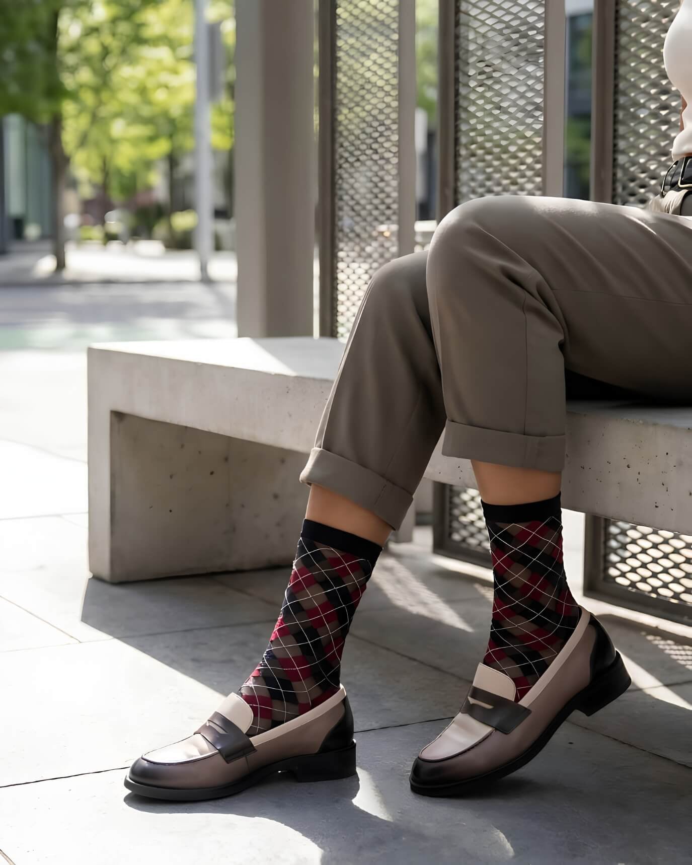 Sheer Argyle Crew Socks in black, red, and beige featuring a classic argyle motif, styled with rolled cuff pants while seated on a sunlit bench, highlighting the semi-sheer material and stylish patterned look.