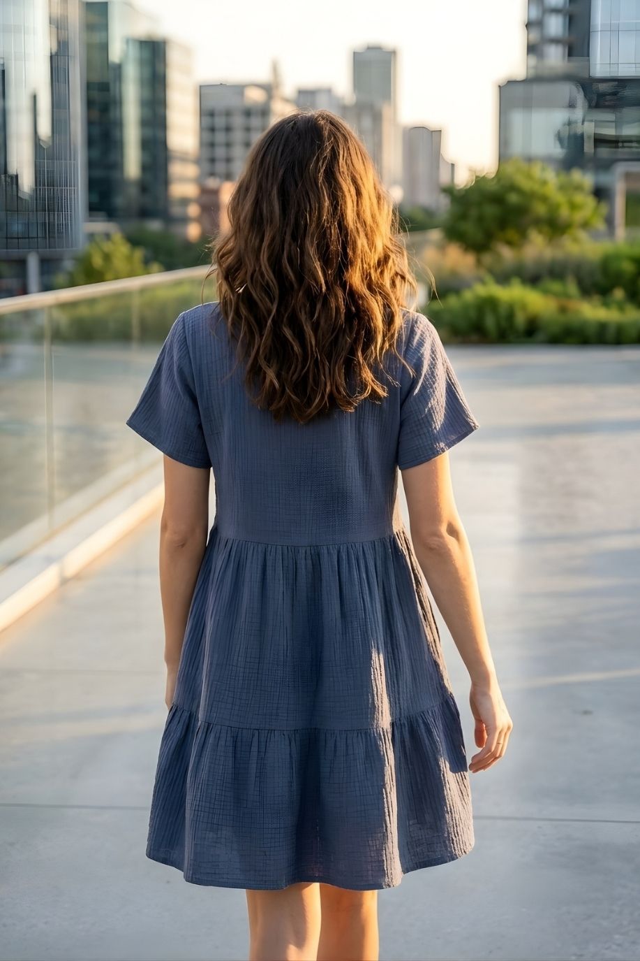 Cassady Dress featuring a loose, comfortable silhouette with a V-neckline, short sleeves, and tiered skirt, shown from the back on a rooftop walkway with greenery and city buildings in the distance.