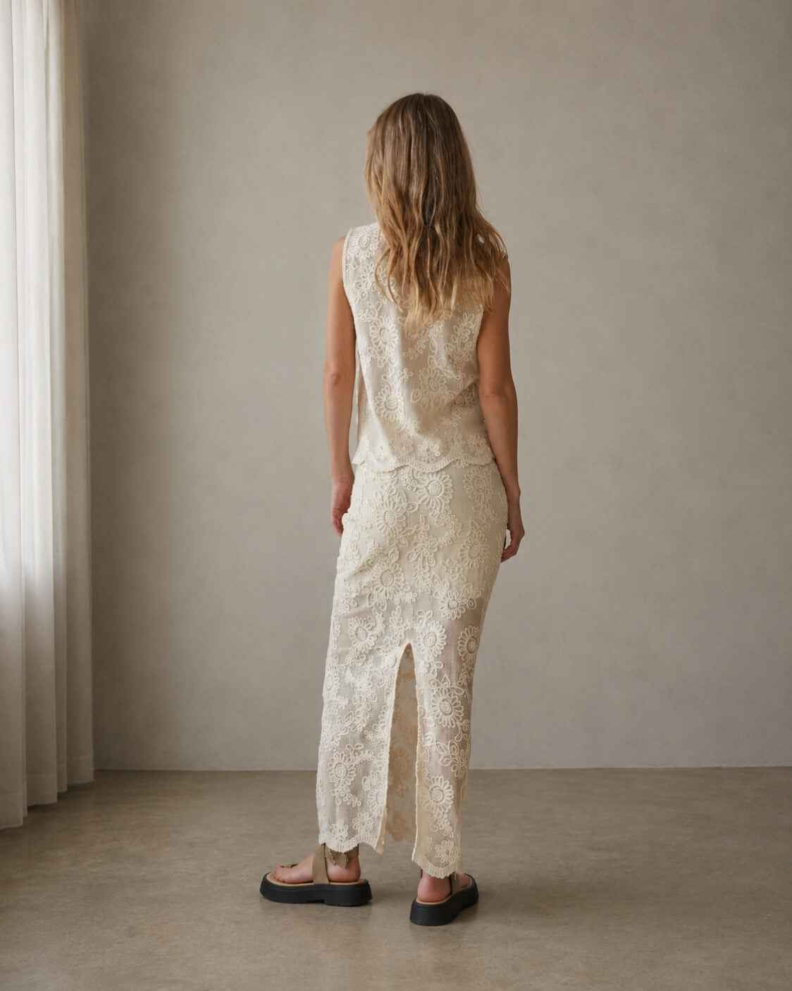 Sleeveless Embroidery Crop Top viewed from the back, featuring intricate floral embroidery across a lightweight sheer mesh fabric with a relaxed drape and scalloped hem, styled with a matching embroidered skirt in a softly lit minimalist interior, highlighting the garment’s detailed craftsmanship from every angle.