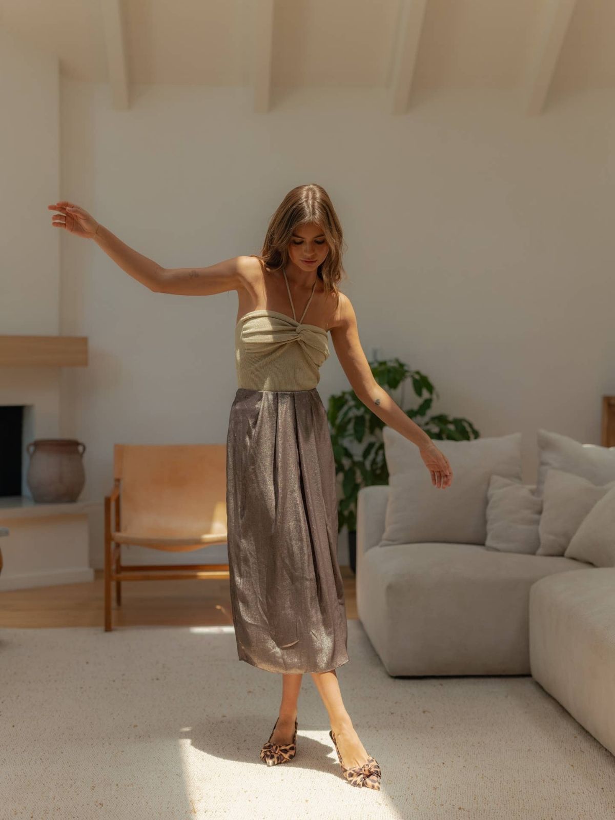 Metallic Fabric Pleat Detailed Skirt, model standing in a bright living room wearing the skirt with a strapless top and heels, highlighting the front pleats and mid length flow.