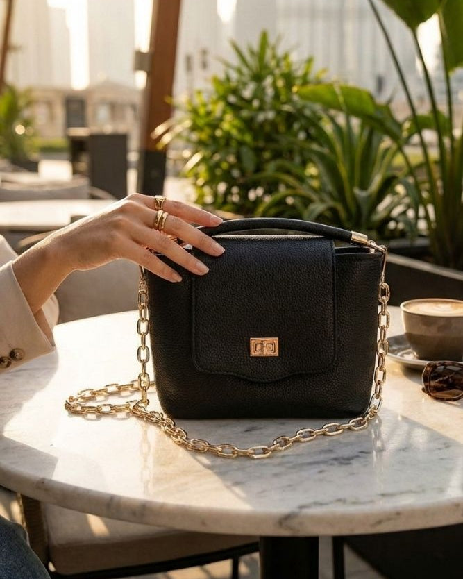 Ismene Leather Purse featuring a pebbled leather body with curved top handle, statement chain strap, and sleek front flap closure, displayed on an outdoor café table in warm sunlight, emphasizing sophisticated detail and modern charm.