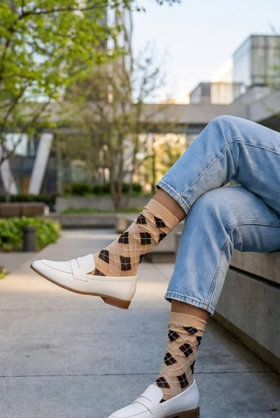 Sheer Argyle Crew Socks in beige and black featuring a soft diamond pattern, paired with white loafers and cuffed jeans while sitting on a concrete bench, showcasing the lightweight, sheer texture and casual styling.
