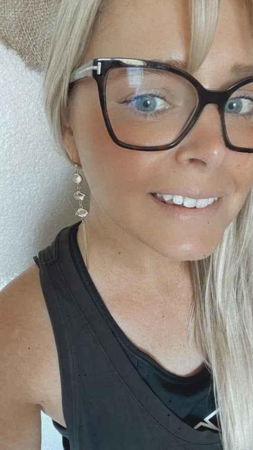 Smiling woman with long blonde hair, wearing large eyeglasses and geometric earrings, black top