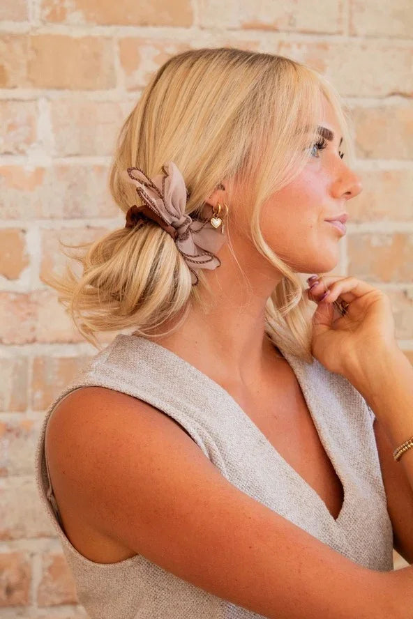 Blonde woman with a taupe hair scrunchie and heart earrings poses against a brick wall, Love Classic style