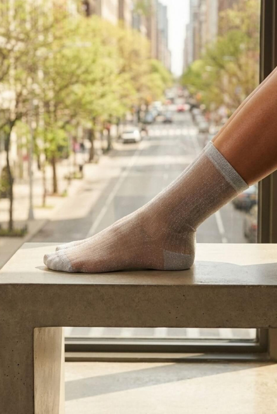 Sheer Metallic Ribbed Crew Socks in sheer silver tone with metallic sheen, displayed on a foot resting on a concrete ledge by a city window, showcasing the transparent lightweight fabric and smooth ribbed construction.