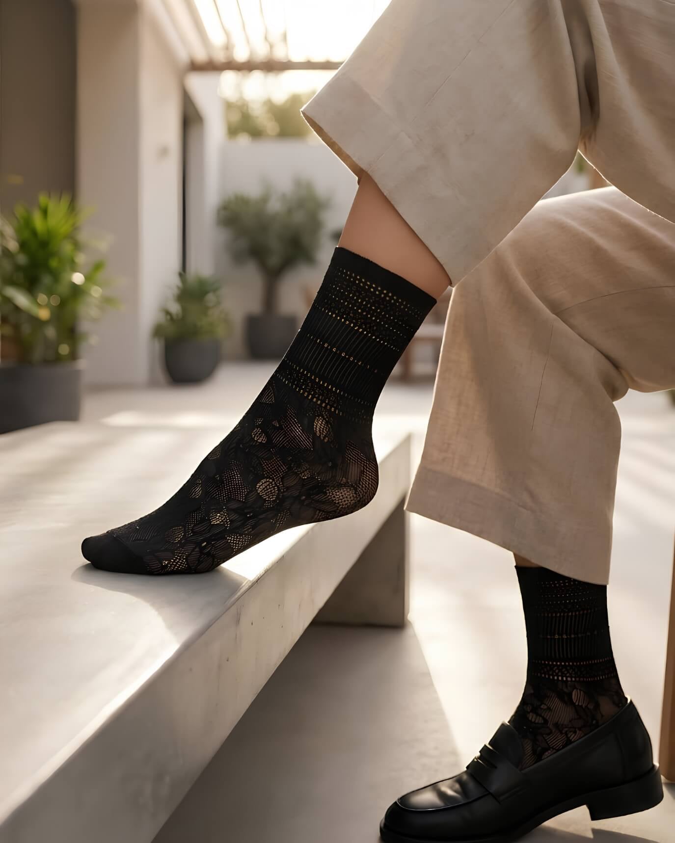 Sheer Floral Lace Crew Socks in black with intricate floral lace design, worn with tailored trousers and black loafers while sitting on a sunlit bench, showcasing the lightweight, sheer finish and elegant pattern.