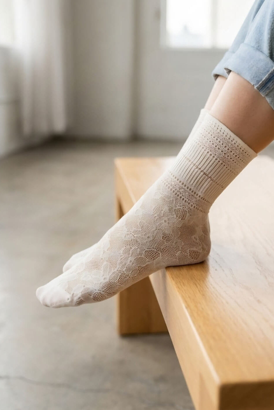 Sheer Floral Lace Crew Socks in ivory with intricate lace floral pattern, worn with rolled cuff jeans while resting on a wooden bench indoors, highlighting the delicate texture and lightweight feel.