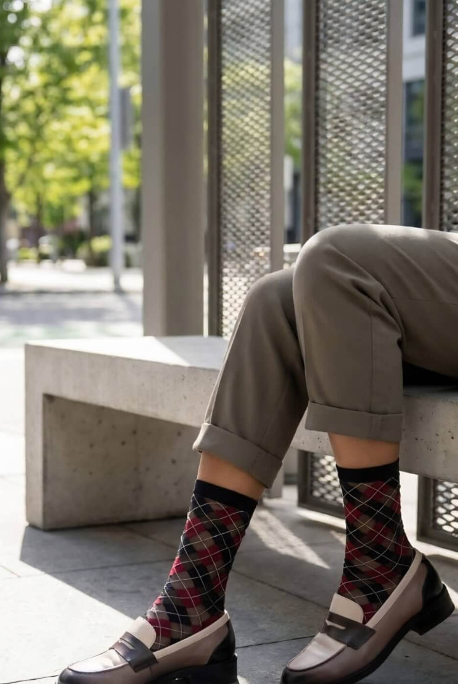 Sheer Argyle Crew Socks in black, red, and beige featuring a classic argyle motif, styled with rolled cuff pants while seated on a sunlit bench, highlighting the semi-sheer material and stylish patterned look.
