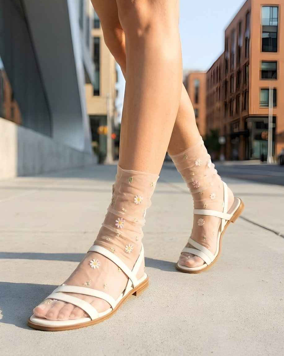 Sheer Garden Daisy Crew Socks in nude sheer fabric with floral embroidery, paired with white strappy sandals while walking on a city sidewalk.