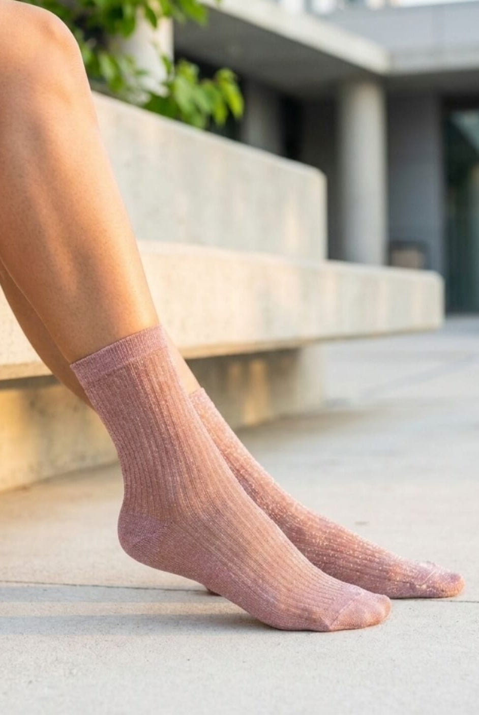Sheer Metallic Ribbed Crew Socks in blush pink with soft shimmer, worn on bare feet while seated on concrete steps, highlighting the flexible ribbed knit and semi-sheer elegant finish.