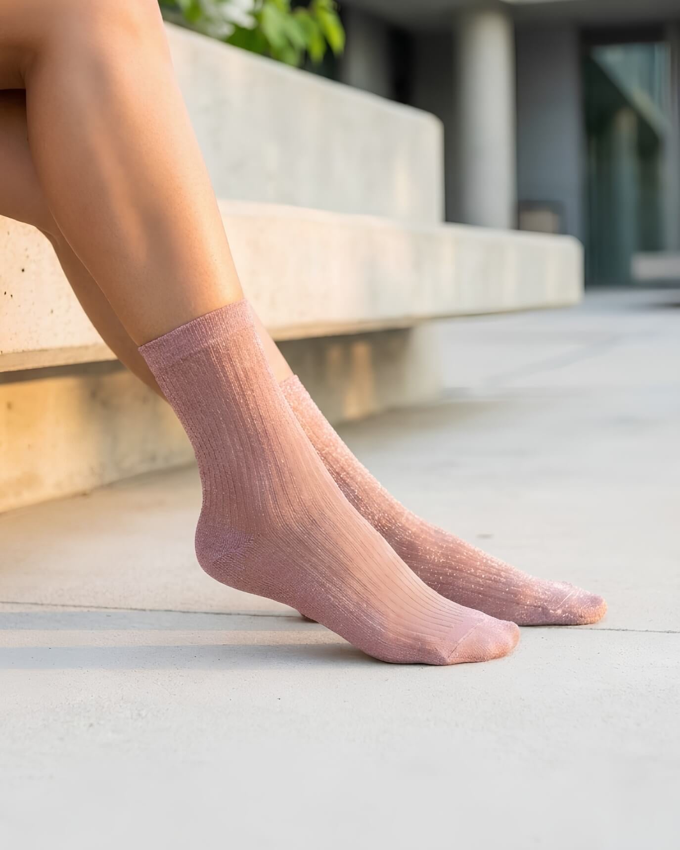 Sheer Metallic Ribbed Crew Socks in blush pink with soft shimmer, worn on bare feet while seated on concrete steps, highlighting the flexible ribbed knit and semi-sheer elegant finish.