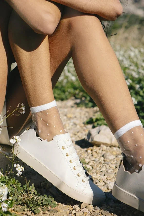 White leather sneakers with polka dot sheer socks outdoors on a gravel path with wildflowers