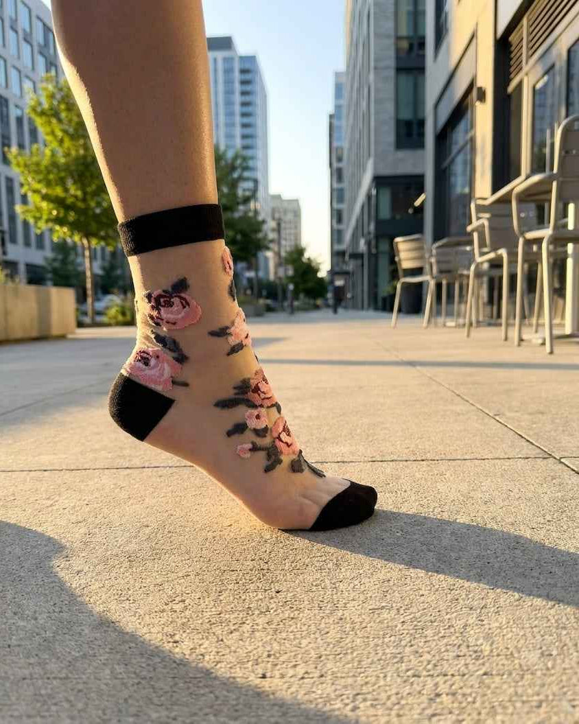 Roses Sheer Socks 2-Pack featuring a black-accent sheer ankle sock with embroidered rose details, worn outdoors on a city walkway in warm sunlight, highlighting the transparent finish and decorative floral embroidery.