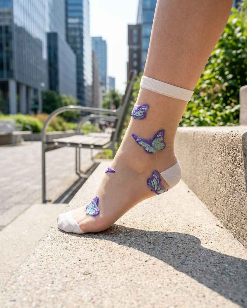 Butterfly Dreams Sheer Socks Bundle Pack featuring a white sheer ankle sock with embroidered butterfly motifs, worn outdoors on a foot resting on a stone ledge in a city setting, highlighting the translucent finish and playful butterfly detailing.