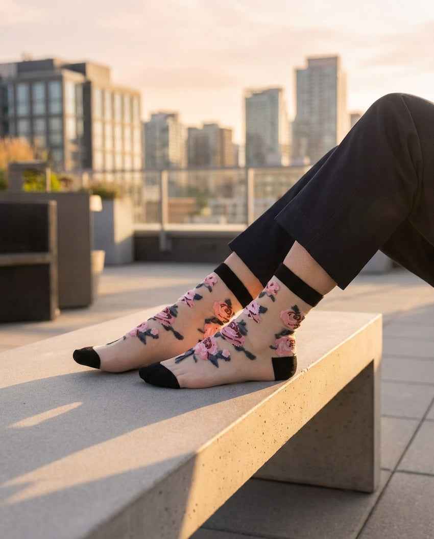 Roses Sheer Socks 2-Pack featuring sheer ankle socks with embroidered rose motifs, black cuff and toe panels, worn while seated on an outdoor bench in a rooftop city setting, highlighting the floral texture and lightweight translucent finish.