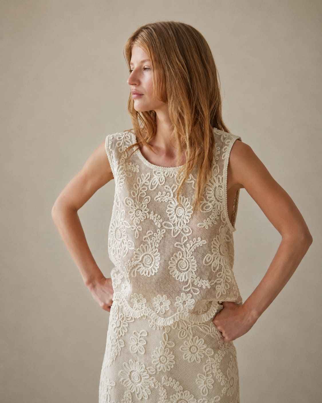 Sleeveless Embroidery Crop Top with ornate floral embroidery and a semi-sheer mesh construction in an elegant ivory shade, styled with a scalloped hem and relaxed sleeveless silhouette, worn in a side-profile pose that showcases the texture and dimensional detailing of the embroidery against a neutral studio backdrop.