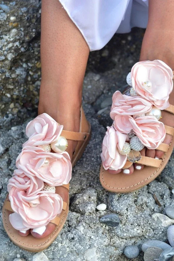 Woman wearing tan sandals with large pink flower decorations and pearls on rocky ground