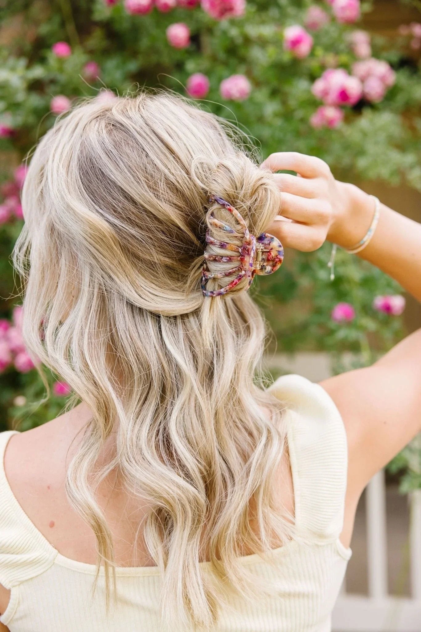 Blonde woman wearing a colorful claw hair clip, styled in loose waves, with pink flowers in background