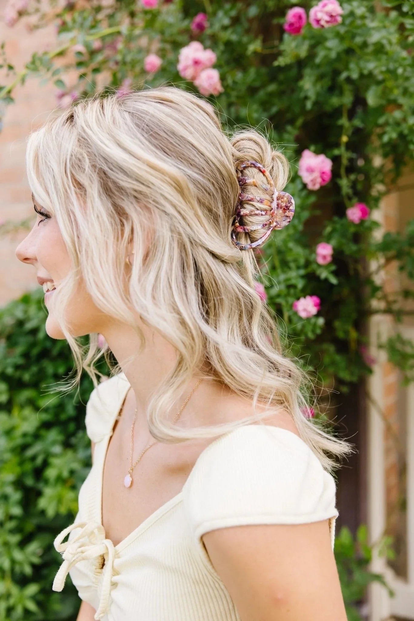Blonde woman wearing a floral claw clip in her hair, outdoors with pink roses, Love Classic style