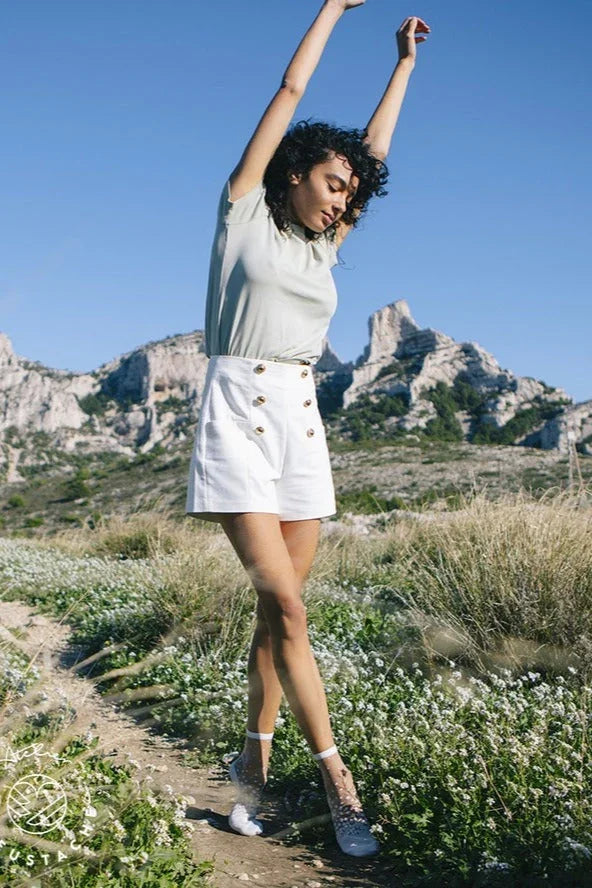 Woman in white high-waisted shorts and light top posing outdoors with mountains in background